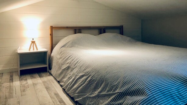 Ein Schlafzimmer mit schrägverdeckter Decke, einem hölzernen Kopfteil, einem Nachttisch mit Lampe und einer gestreiften Bettdecke.