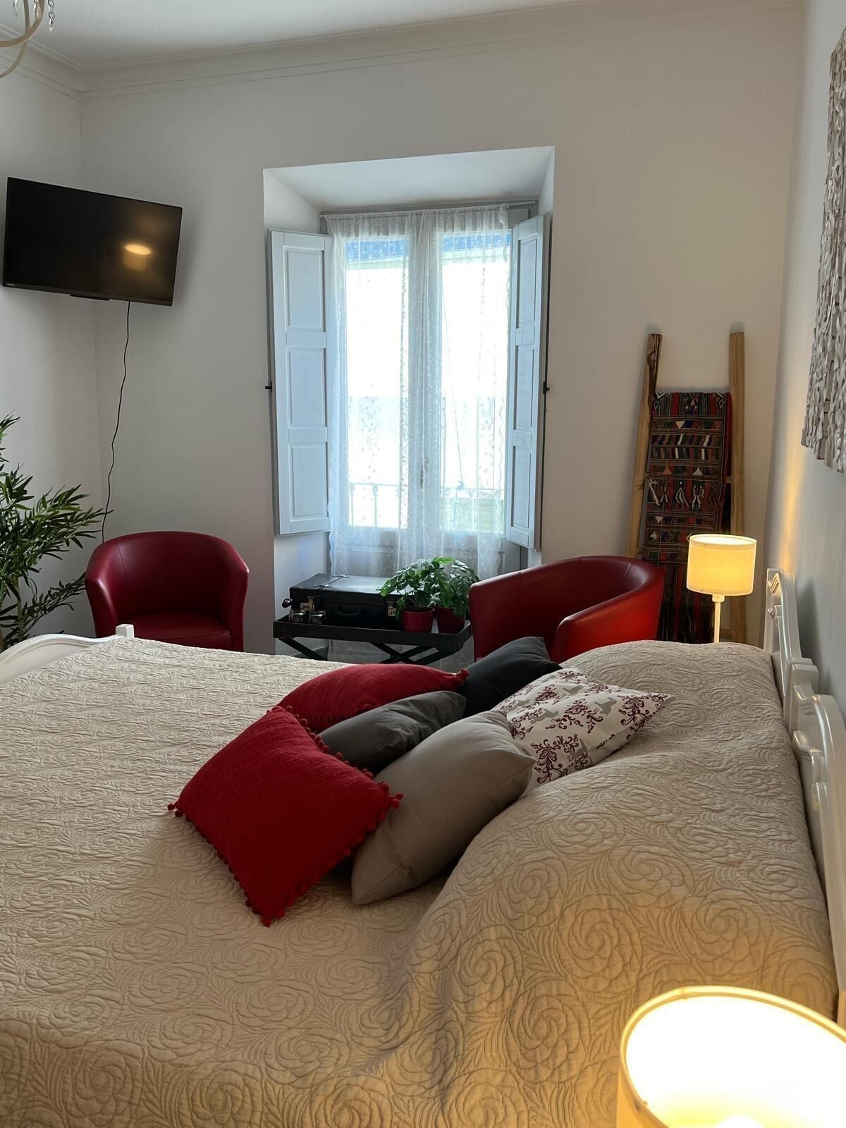 Une chambre avec un grand lit, deux fauteuils rouges, une petite table avec une plante et un téléviseur fixé au mur.
