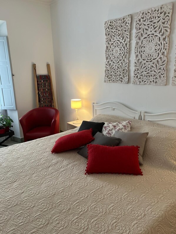 A bedroom with a bed, red and grey pillows, a red chair, a nightstand with a lamp, and decorative wall art.
