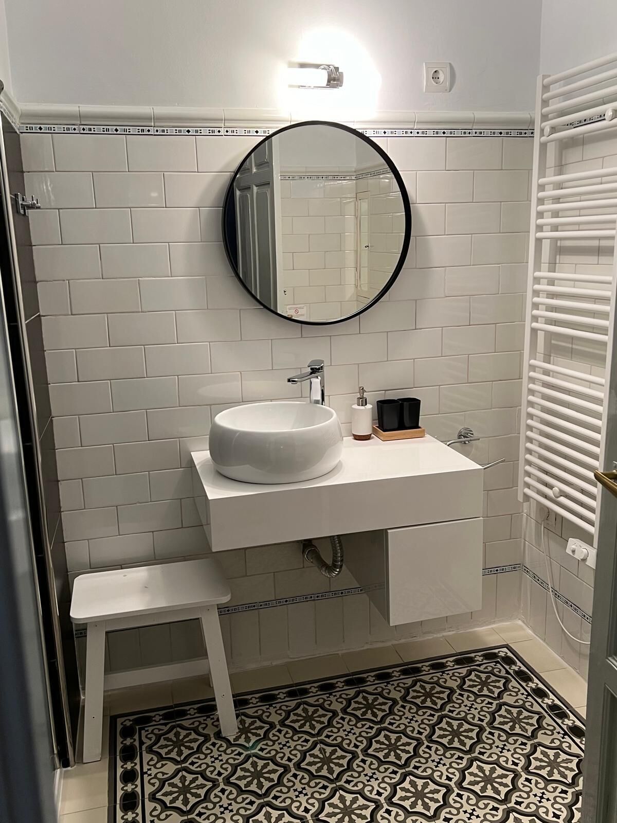 A bathroom with white subway tiles, a round mirror, a white sink, a small stool, and a patterned floor.