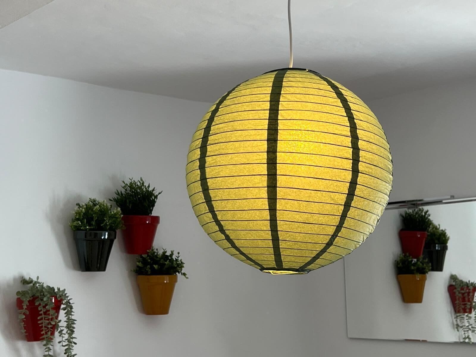 Una lámpara colgante esférica con un diseño a rayas amarillo y negro, rodeada de plantas en macetas sobre una pared blanca.