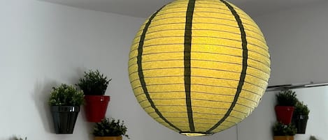 Una lámpara colgante esférica con un diseño a rayas amarillo y negro, rodeada de plantas en macetas sobre una pared blanca.