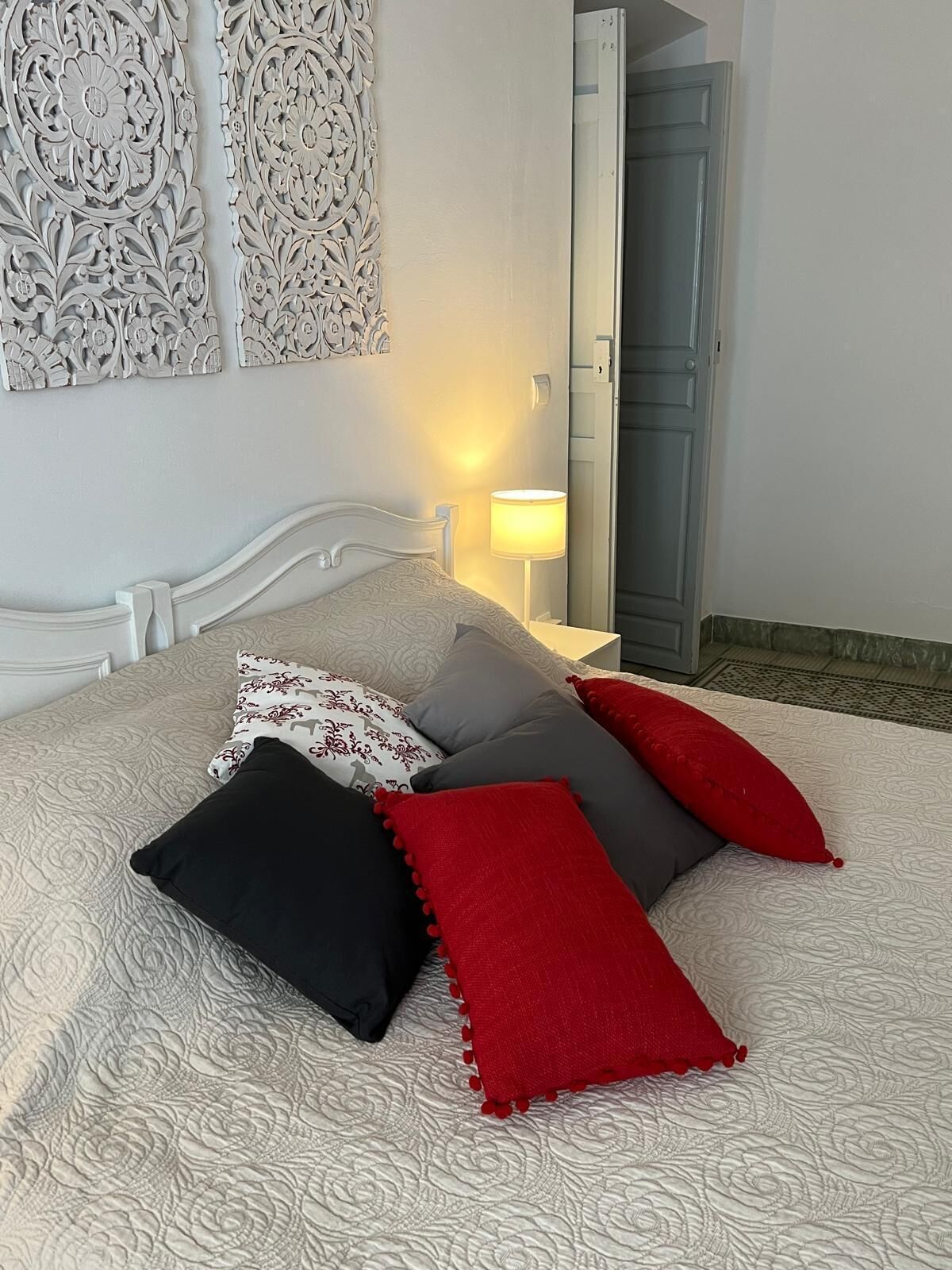 Una cama bien hecha con ropa de cama estampada y cojines decorativos, una lámpara de noche y una puerta que da a otra habitación.