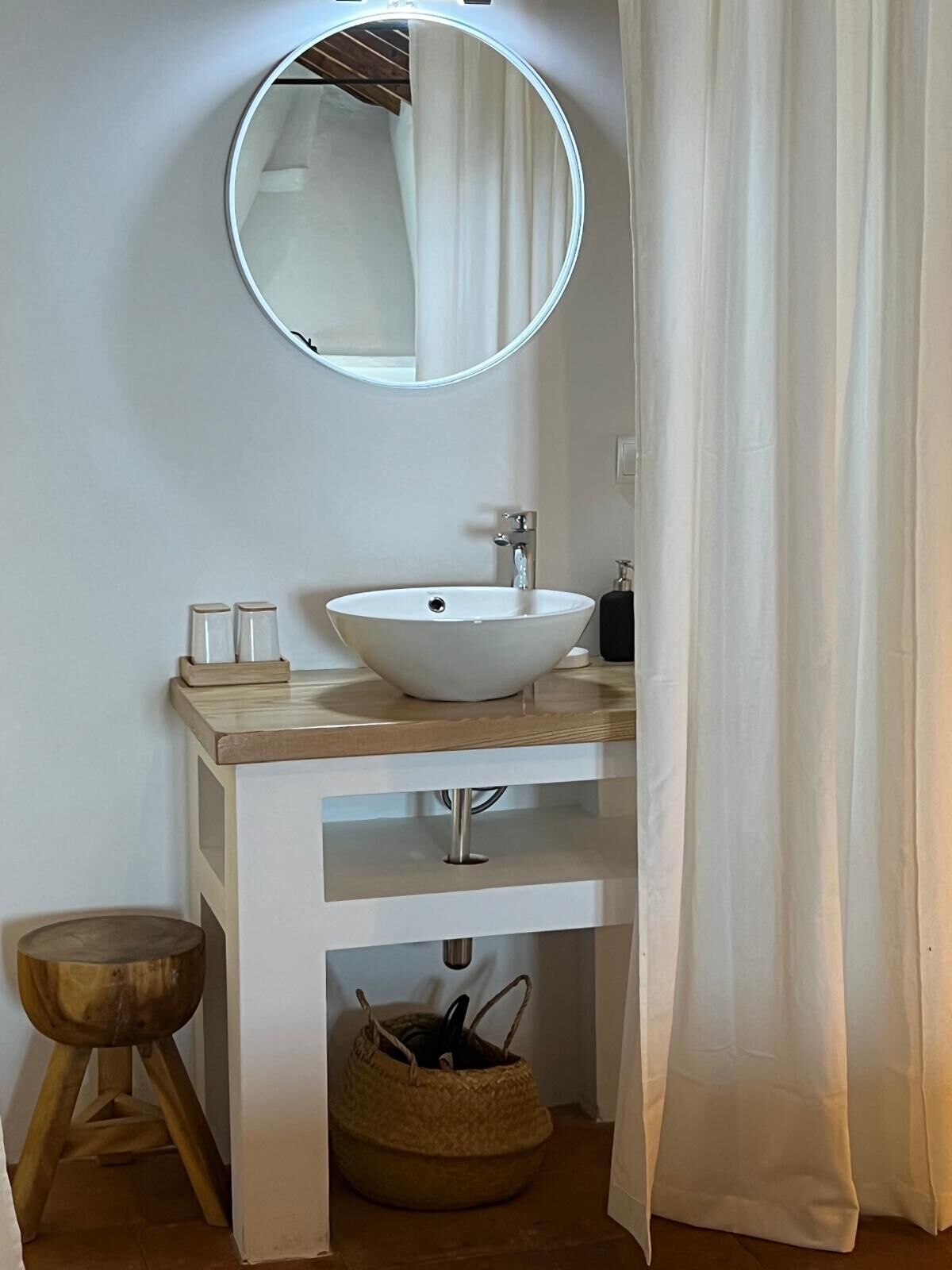 A bathroom with a round mirror, a white sink, a wooden stool, and a woven basket.