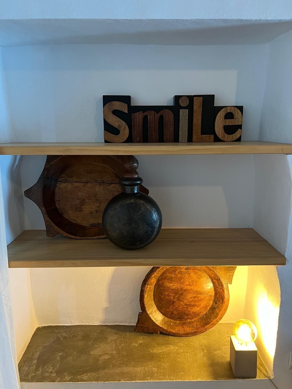 Una estantería con un cuenco de madera, un florero oscuro y un bloque de madera que deletrea "Smile".