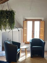 Una habitación con dos sillones azul oscuro, una mesita redonda, una planta colgante y una puerta de madera con contraventanas.