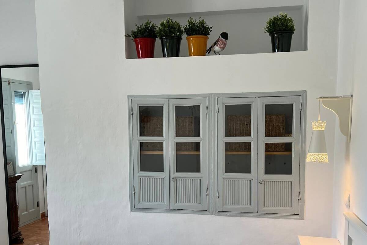 A bedroom with a bed, pillows, a nightstand, a wall-mounted shelf with potted plants, and a window with shutters.