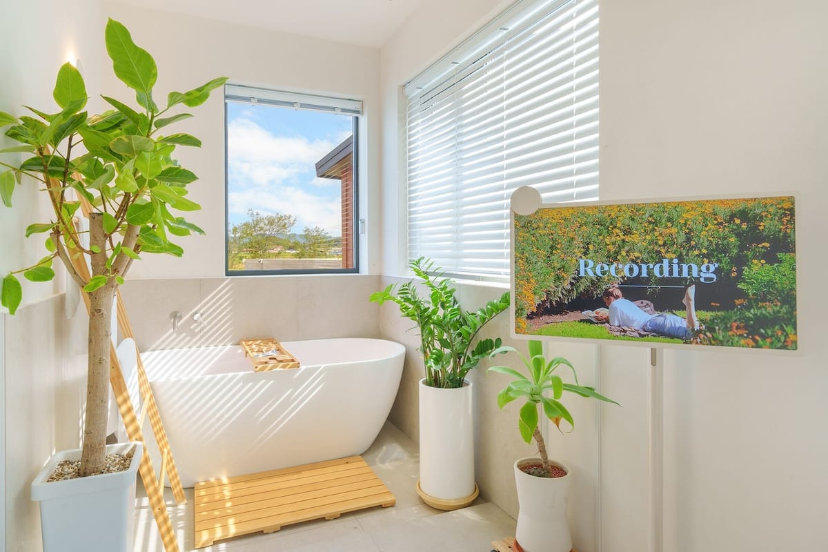 A modern bathroom with a freestanding bathtub, potted plants, and a window with blinds.
