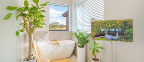 A modern bathroom with a freestanding bathtub, potted plants, and a window with blinds.