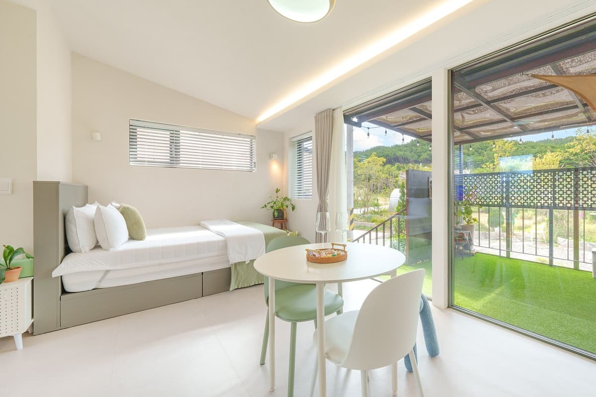 A modern room with a bed, a dining table with chairs, a balcony with a view of greenery and a fence.