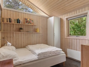 A bedroom with a bed, a wardrobe, a shelf with books and a painting, and a window with a view of trees.