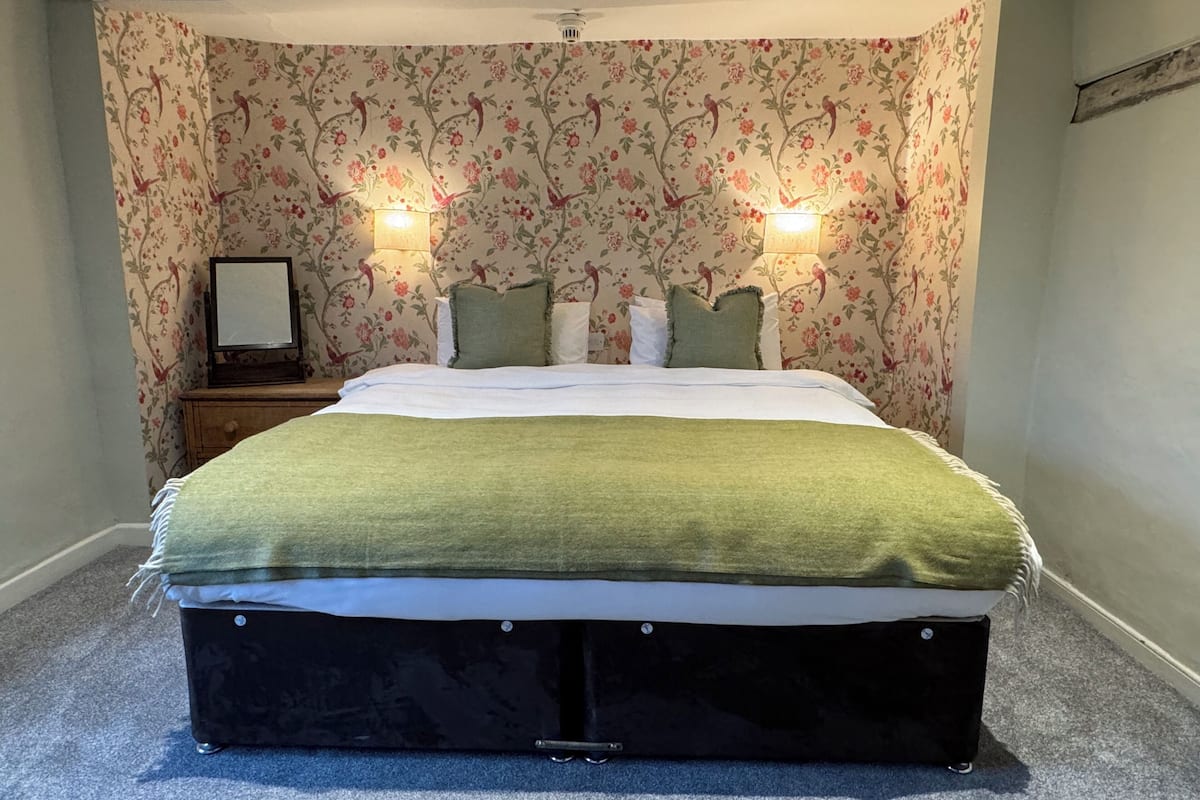 Un dormitorio con papel tapiz floral, una cama con manta verde, dos lámparas de noche y una mesita de noche con un cuadro enmarcado.