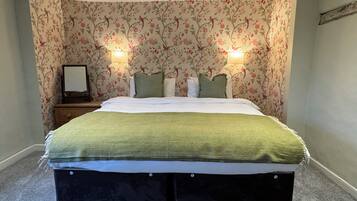 A bedroom with a floral wallpaper, a bed with a green blanket, two bedside lamps, and a nightstand with a framed picture.