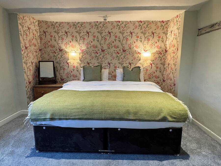 A bedroom with a floral wallpaper, a bed with a green blanket, two bedside lamps, and a nightstand with a framed picture.