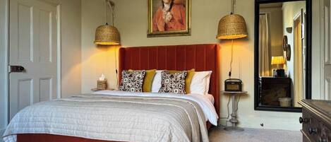 A bedroom with a red upholstered headboard, a large bed with a quilted white bedspread, two bedside lamps, a radio, and a framed painting above the bed.