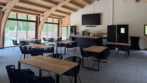 A spacious indoor area with high wooden ceilings, multiple tables and chairs, a bar area with a flat-screen TV, and large windows offering views of the outdoors. - Le moulin de barette (Blavozy)