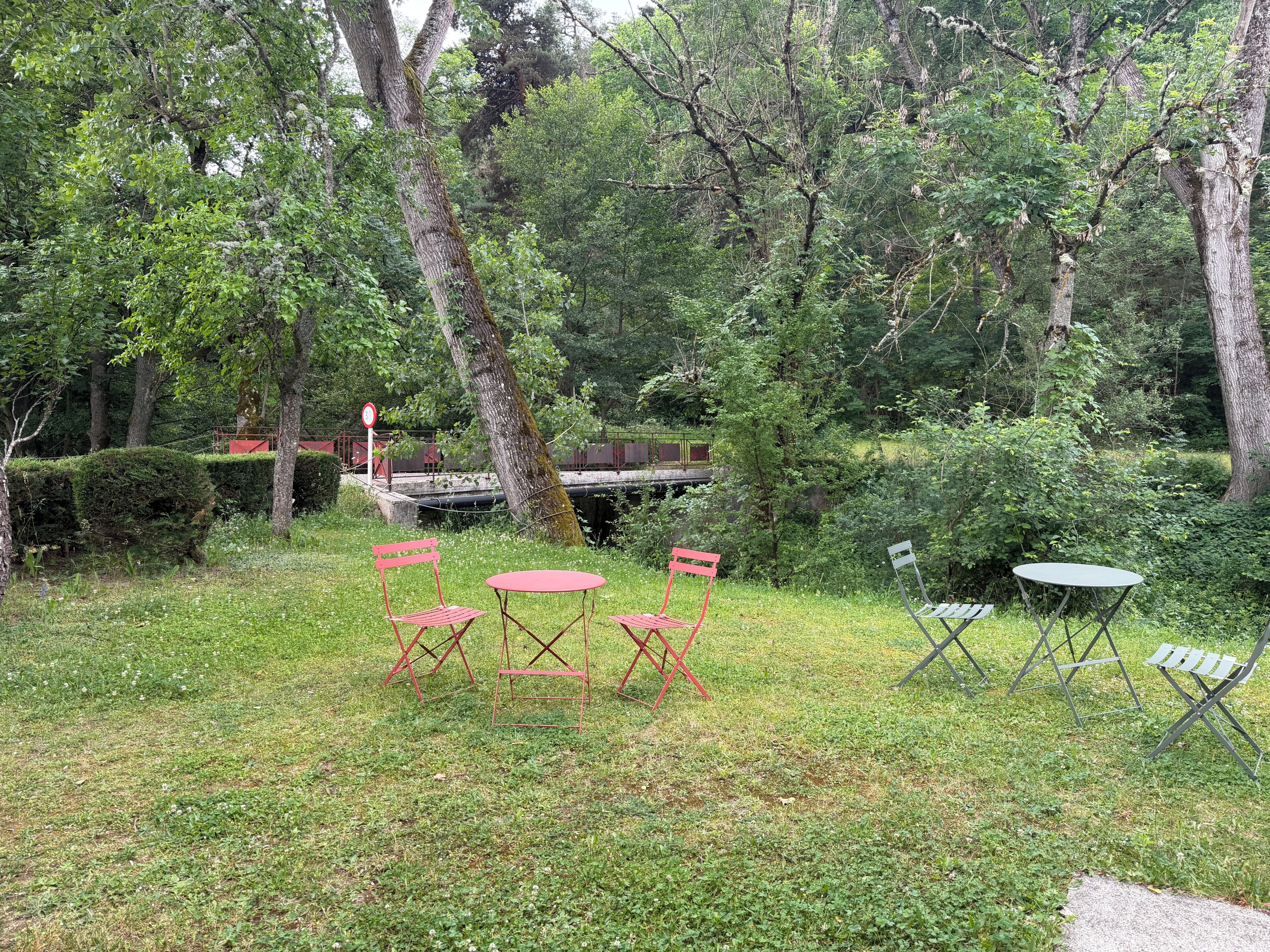 Et græsbevokset udendørsområde med et picnicbord og stole, omgivet af træer og en bro i baggrunden.