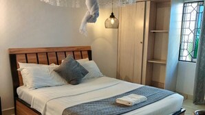 A neatly made bed with a wooden headboard, a bedside lamp, and a window with a view of greenery. - Chamiachi Hotel and Luxury Apartments  (Mombasa)
