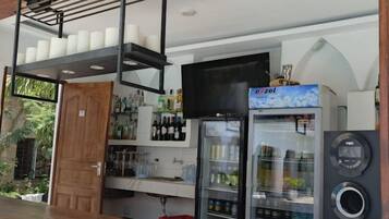 Uma área de bar com balcão de madeira, uma TV fixada na parede, geladeira abastecida com bebidas e sistema de som.