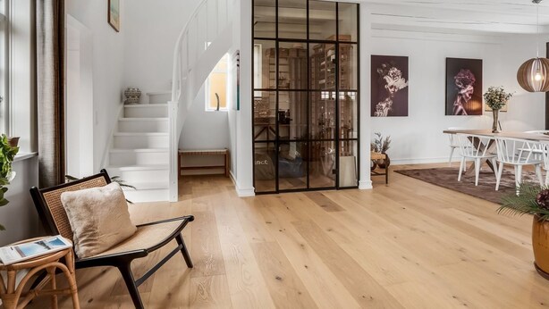 Ein geräumiger Wohnbereich mit Holzboden, einer Treppe, einem Essbereich mit weißem Tisch und Stühlen sowie einer großen Glaswand.
