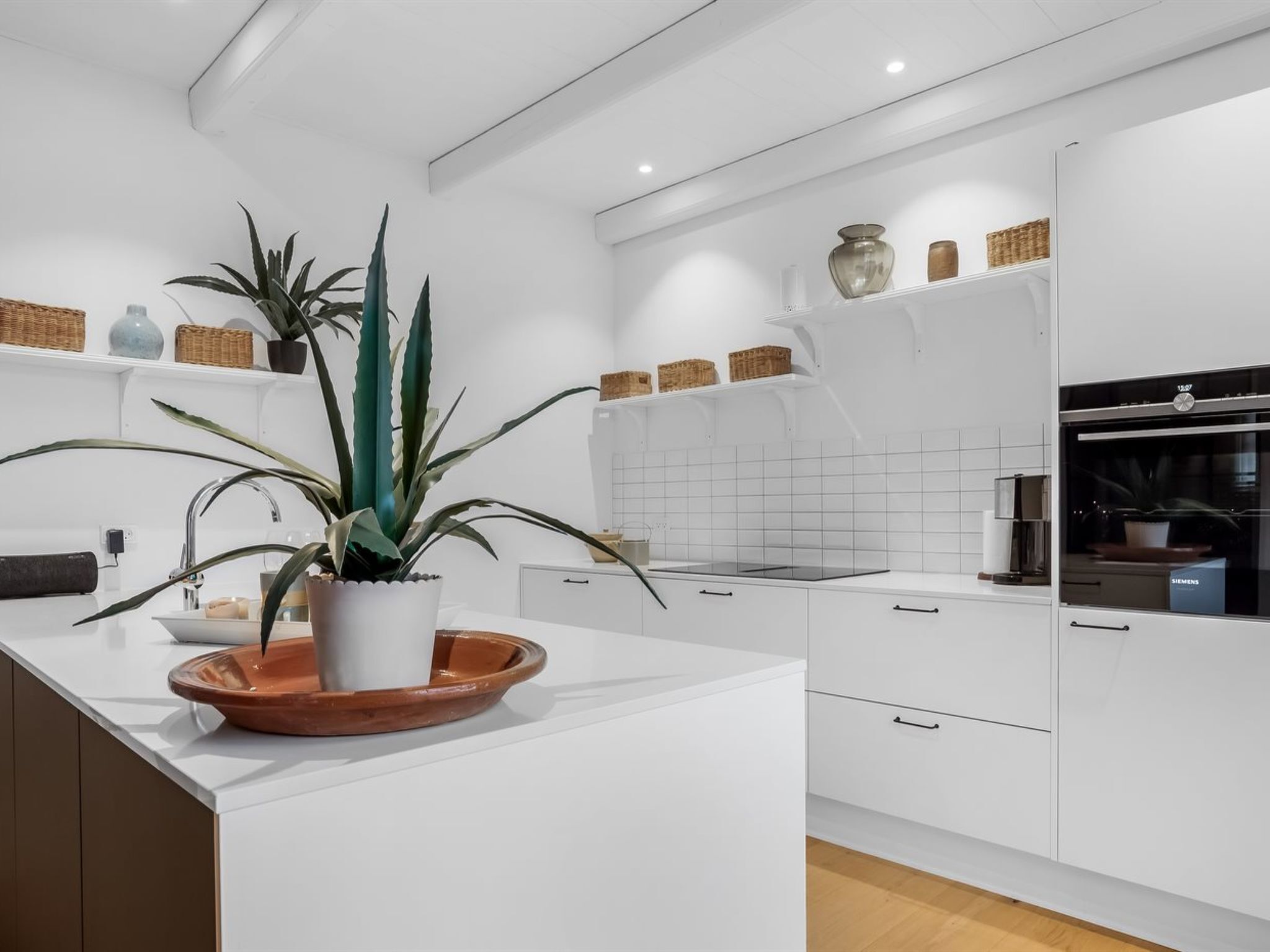 Une cuisine moderne avec des meubles blancs, un îlot central agrémenté d’une plante en pot et un four encastré.