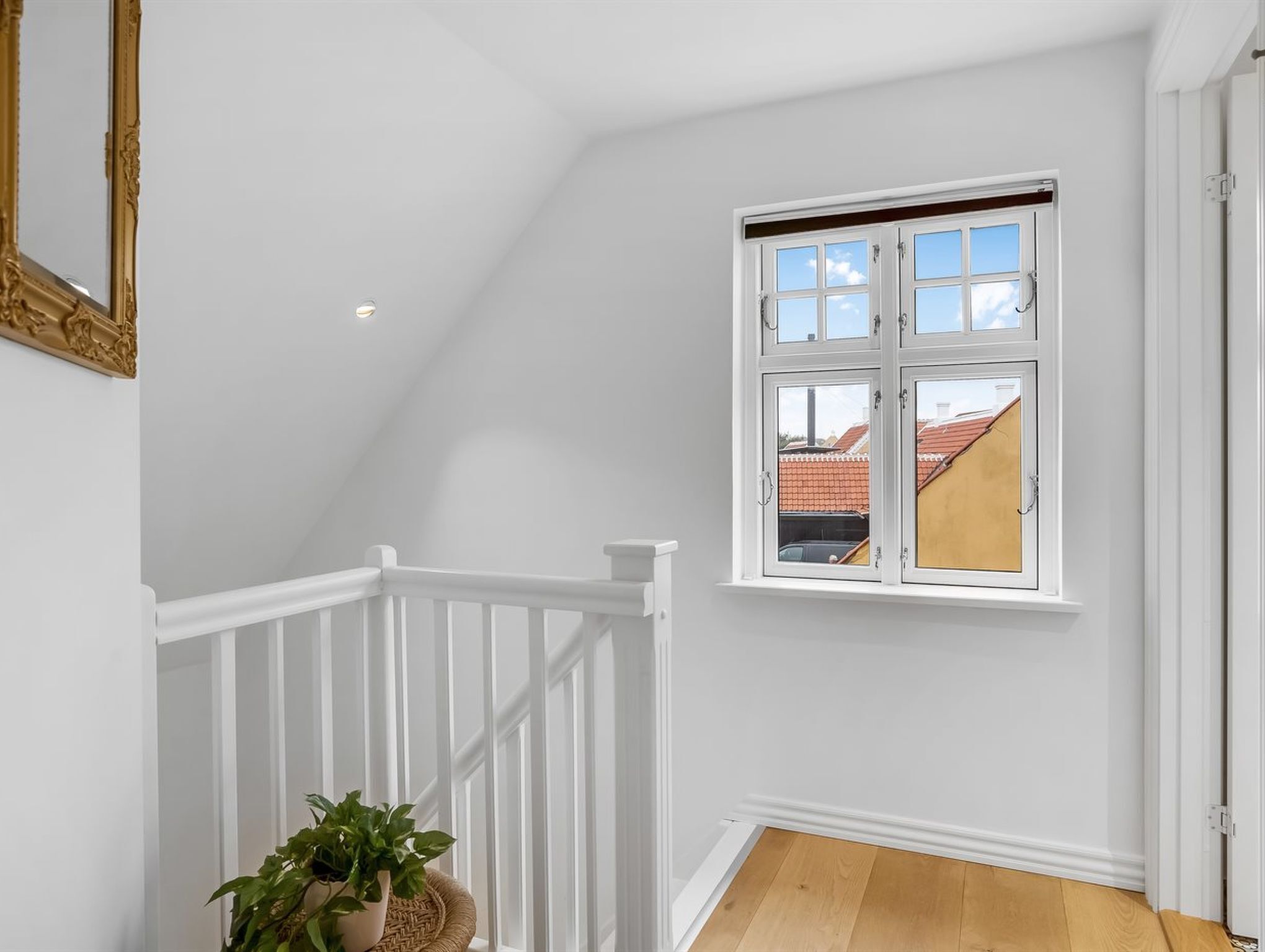 Una habitación con techo blanco, barandilla blanca, una planta en maceta y ventana con vistas a edificios.