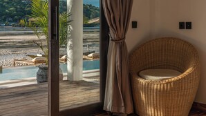 A room with a large sliding glass door leading to a beach view, a wicker chair, and a wooden floor. - La Villa Ceningan by K-Club (Ceningan Island)