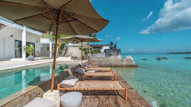 A wooden deck with lounge chairs and umbrellas overlooking a clear blue pool and turquoise sea. - La Villa Ceningan by K-Club (Ceningan Island)