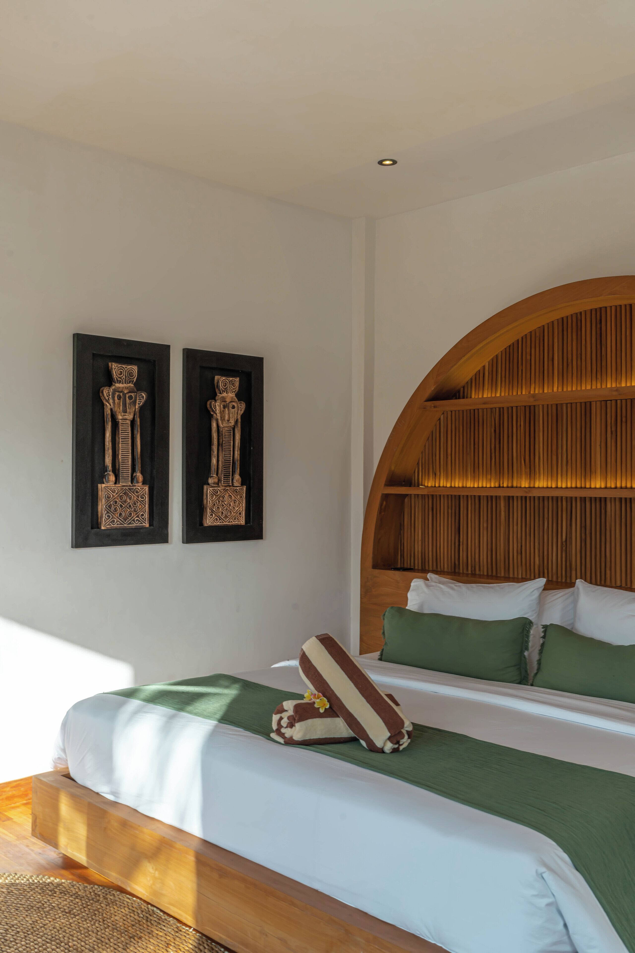 A bedroom with a wooden headboard, two wall-mounted sculptures, a bed with green bedding, and a woven mat at the foot of the bed.