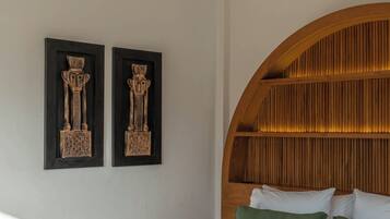 A bedroom with a wooden headboard, two wall-mounted sculptures, a bed with green bedding, and a woven mat at the foot of the bed.