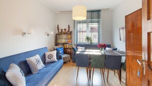 Una sala de estar con un sofá azul, una mesa de comedor con sillas, una estantería y un ventanal con cortinas.