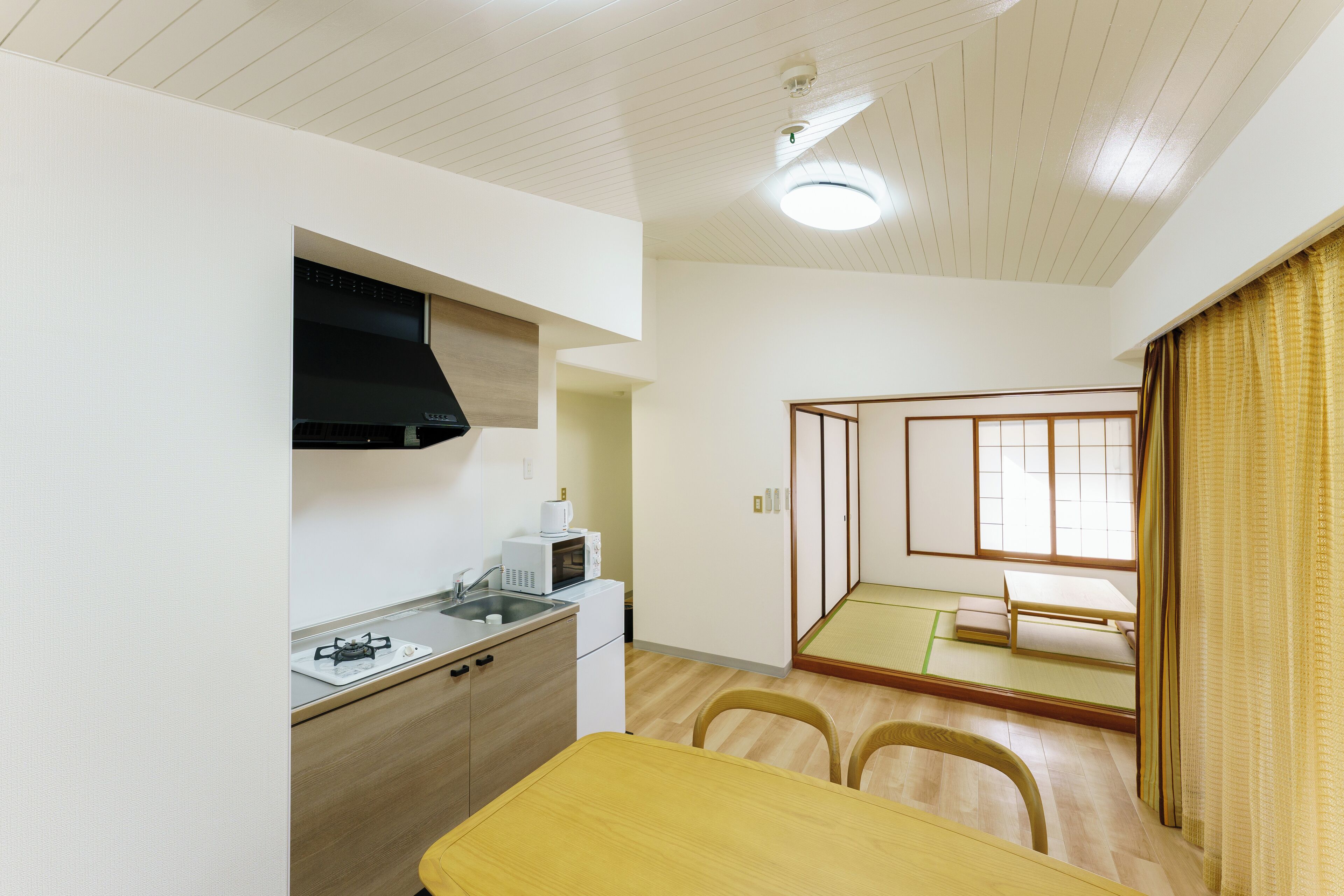 Una cocina compacta con estufa a gas, microondas y una campana extractora negra. En la habitación contigua, hay una mesa de comedor con sillas y un futón de tatami.