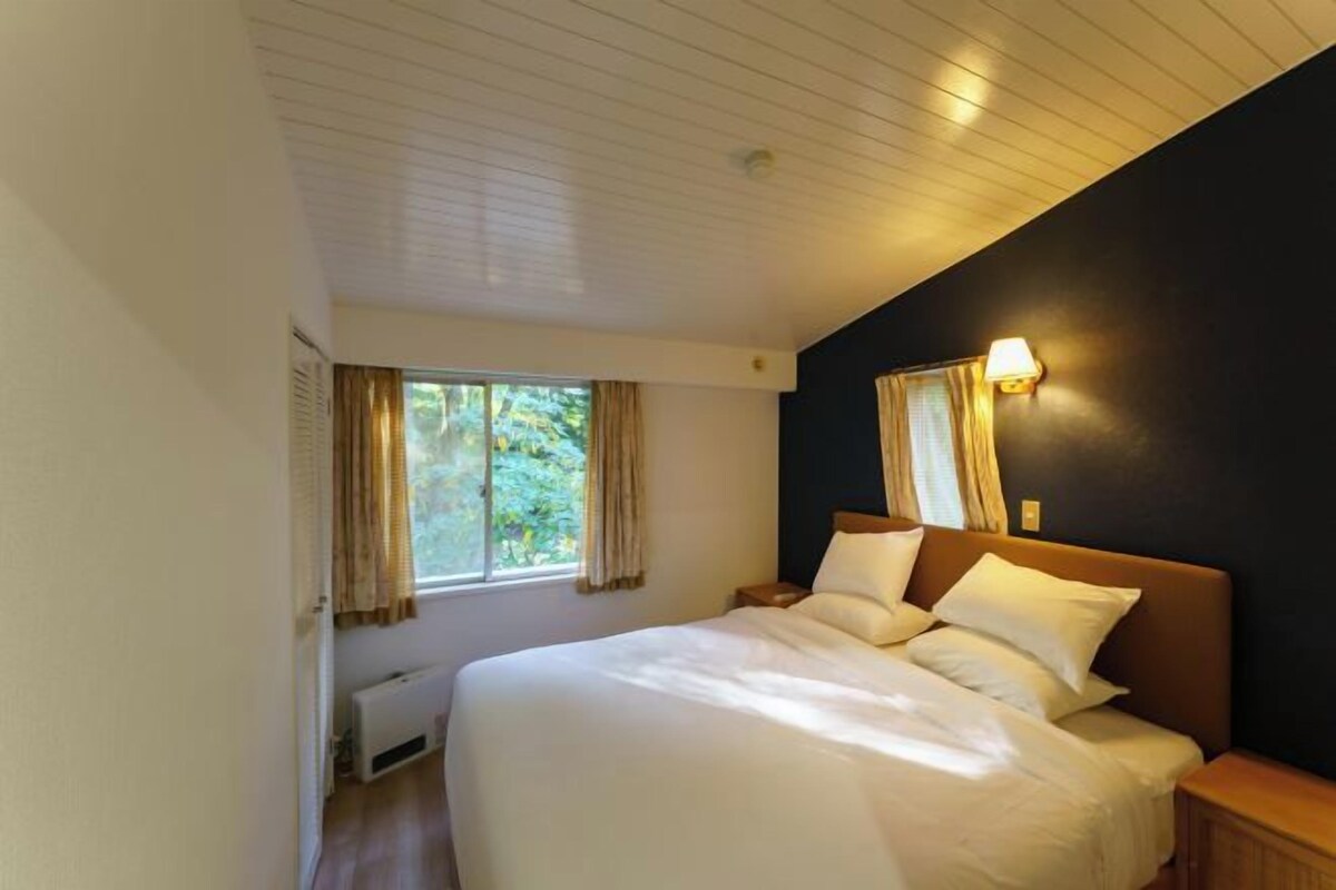 A bedroom with a large bed, a window with curtains, a wall-mounted lamp, and a radiator.