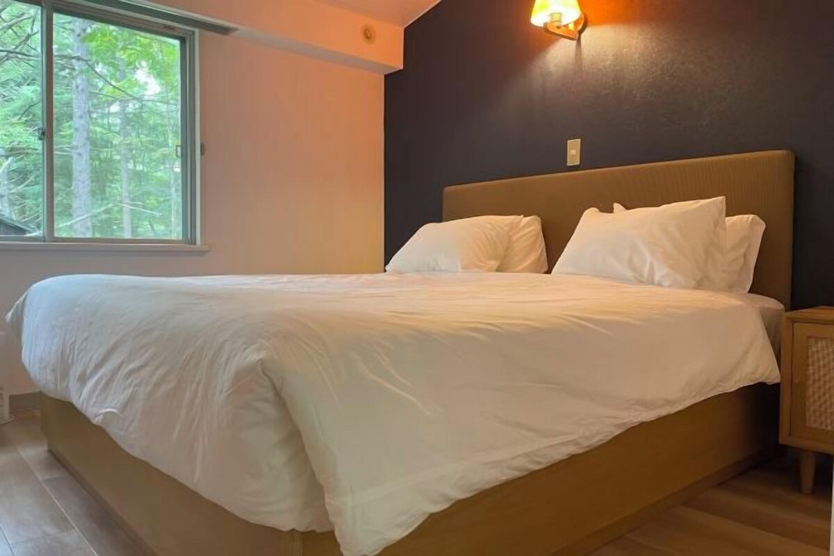 A neatly made bed with white linens in a room with a dark wall and a window with a view of trees.