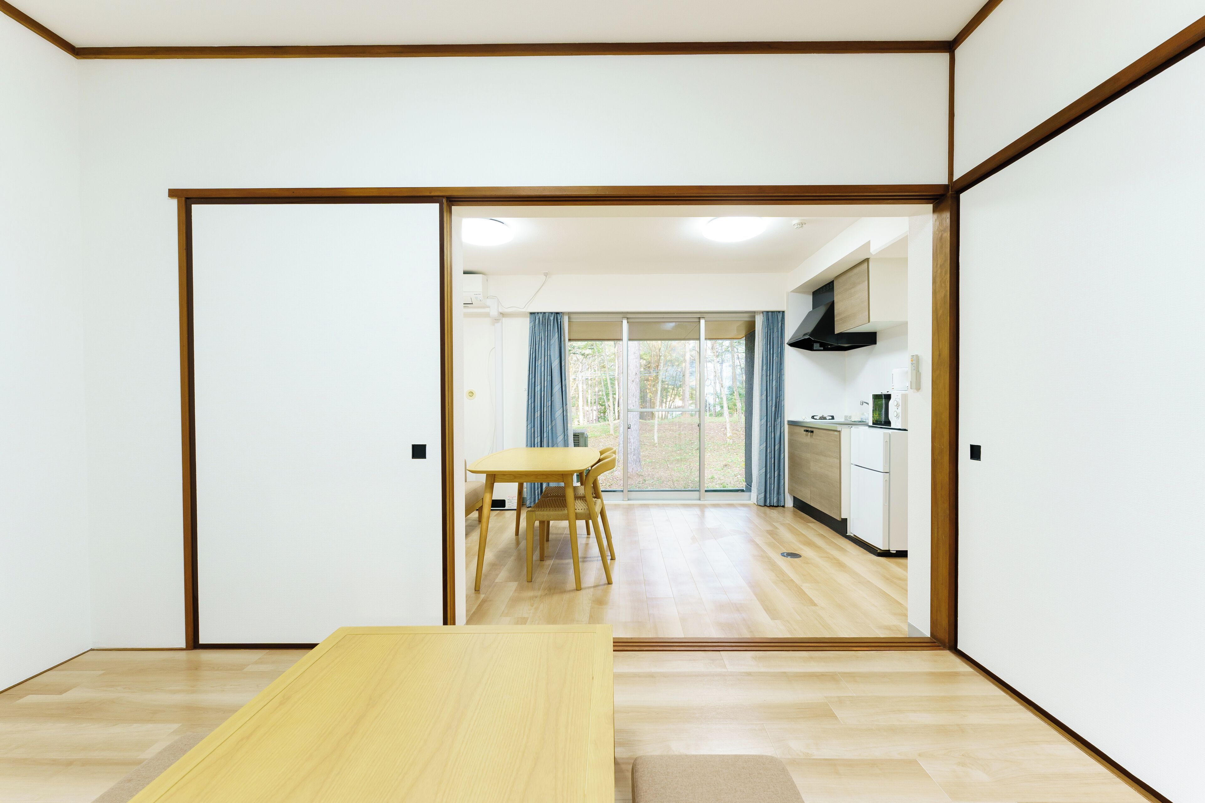 A room with a wooden floor, a dining table with chairs, and a sliding door leading to a kitchen area with a counter and cabinets.