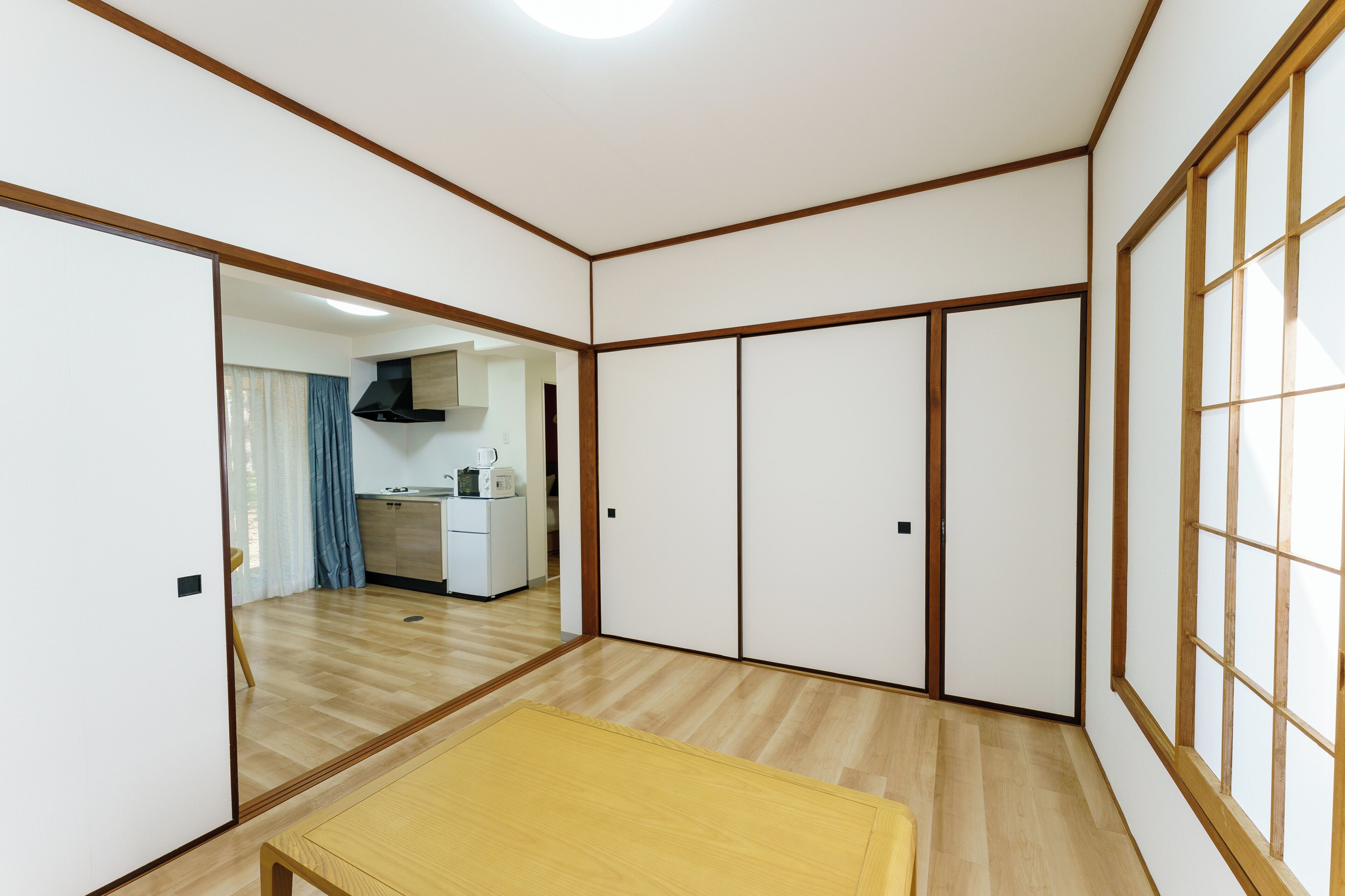 A room with a wooden floor, sliding doors, a kitchen in the background, and a small table.