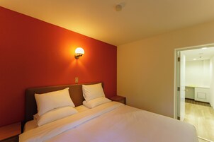 Quarto de hotel com uma cama, mesa de cabeceira, luminária na parede e porta para um banheiro.