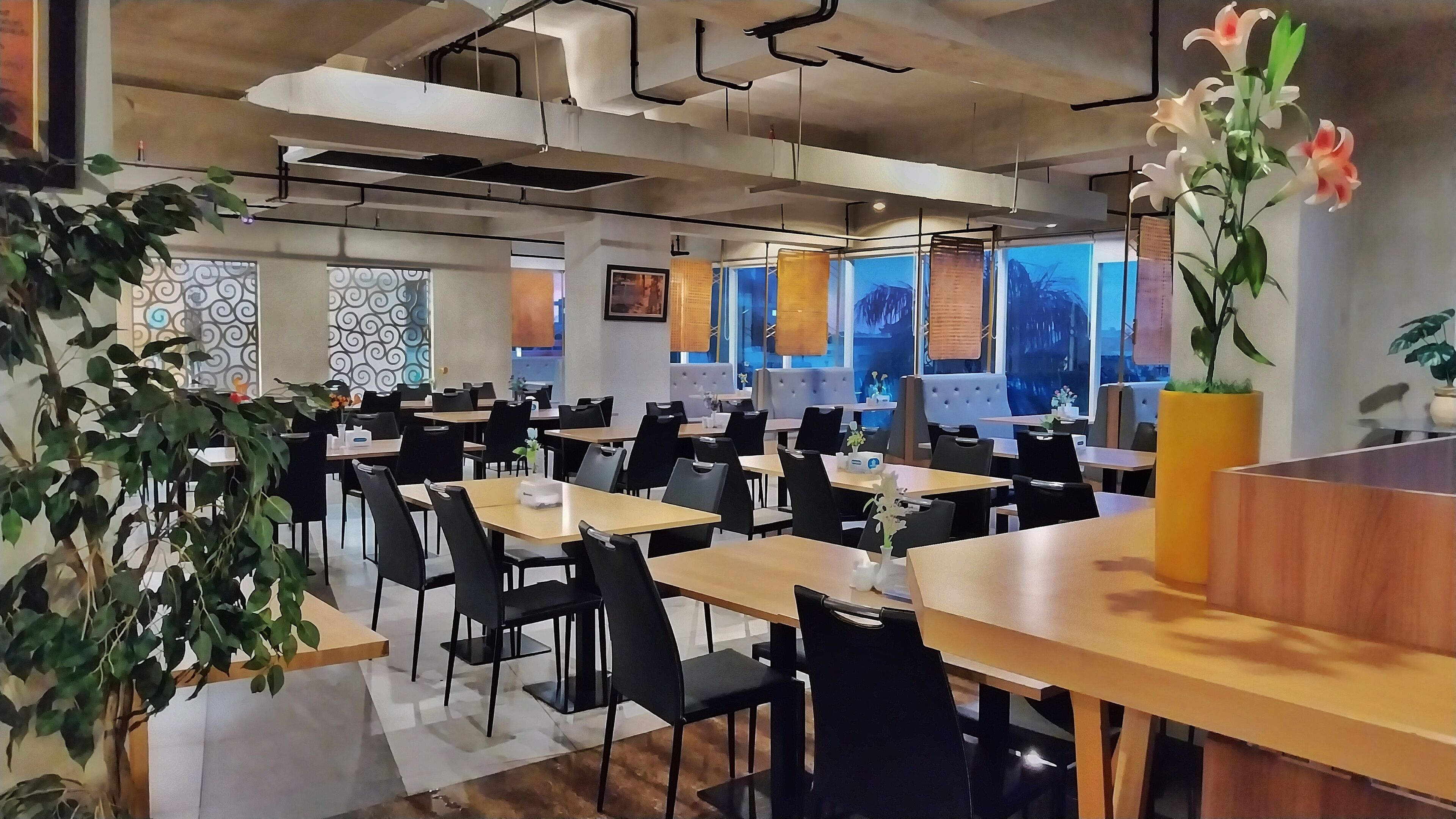 Un restaurante moderno con varias mesas y sillas, ventanas amplias y plantas decorativas.