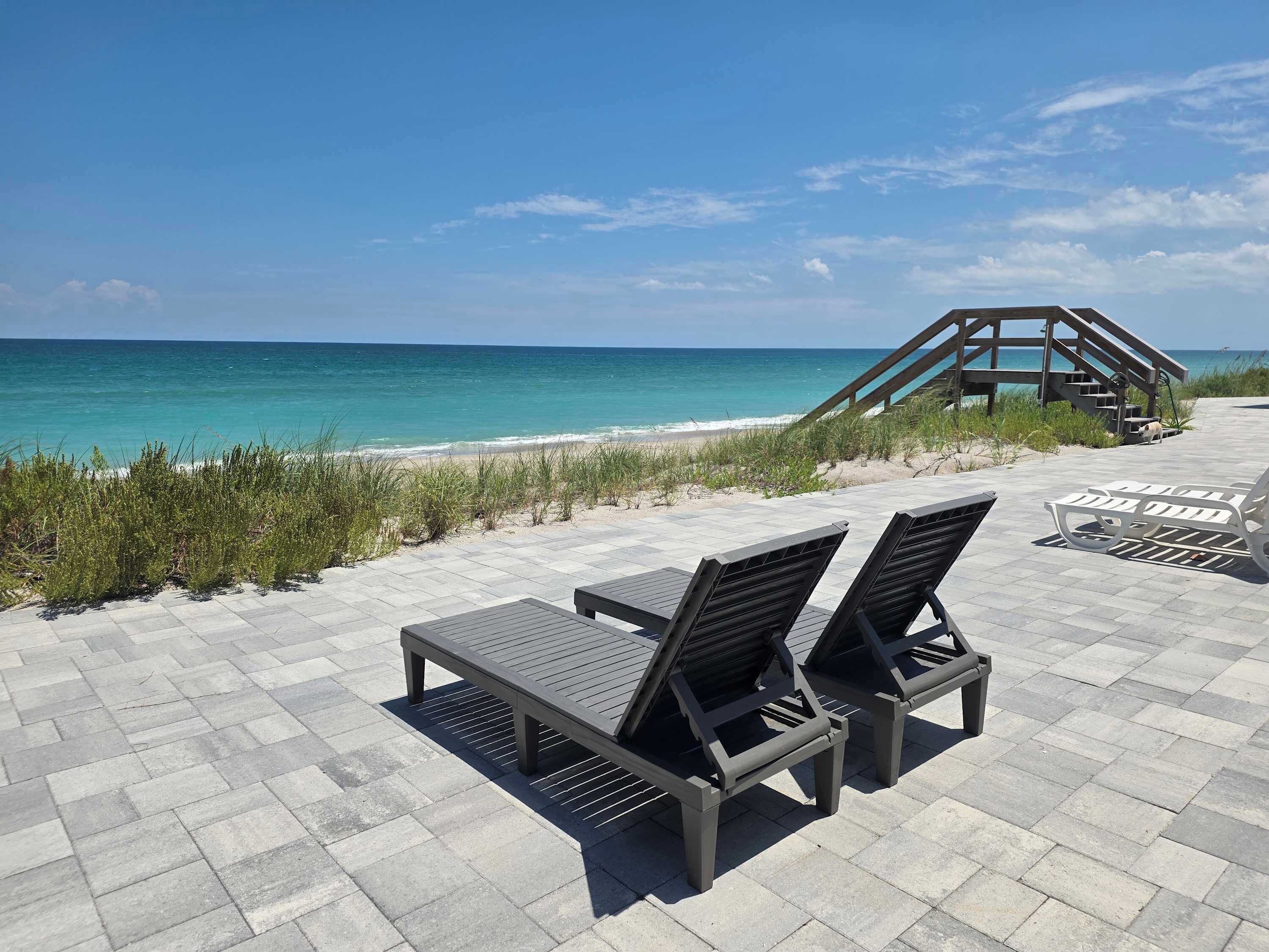 A beachside patio with lounge chairs and a wooden staircase leading to a beach.
