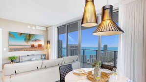 A modern living room with a large window, a sofa, a dining table set for two, and a city view. - 3BR Apartment with Views and Complementary Parking (Hollywood)