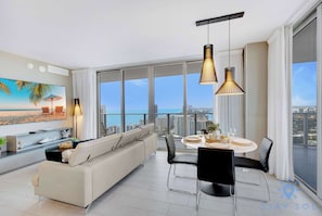 A modern living room with a sofa, dining table, and chairs, large windows with a city view, and pendant lighting. - 3BR Apartment with Views and Complementary Parking (Hollywood)
