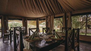 Uma área de refeições com uma mesa posta para quatro pessoas, cercada por cadeiras de madeira, sob uma grande estrutura tipo tenda com janelas.