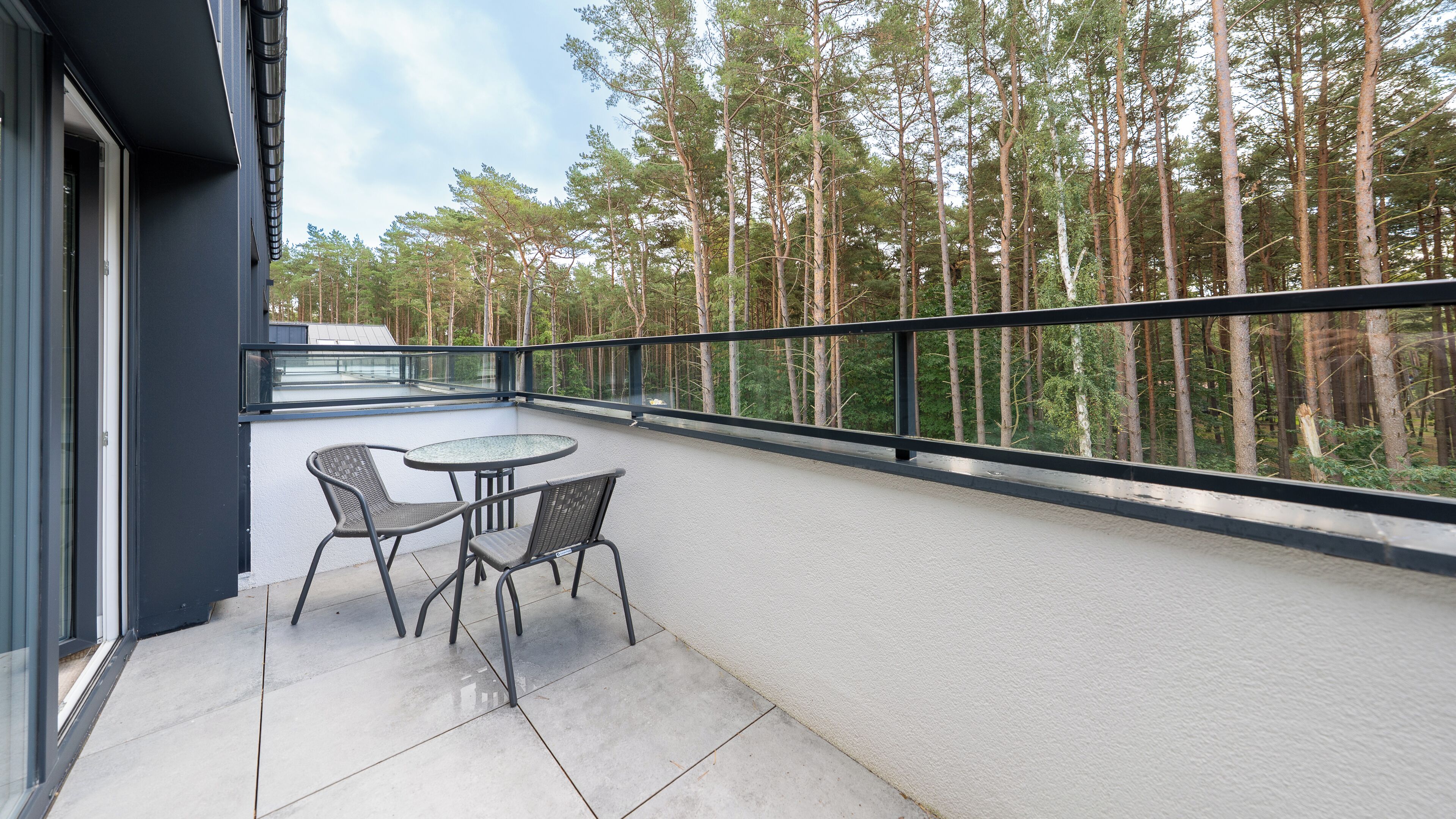 A balcony with a glass table and chairs, overlooking a forest.