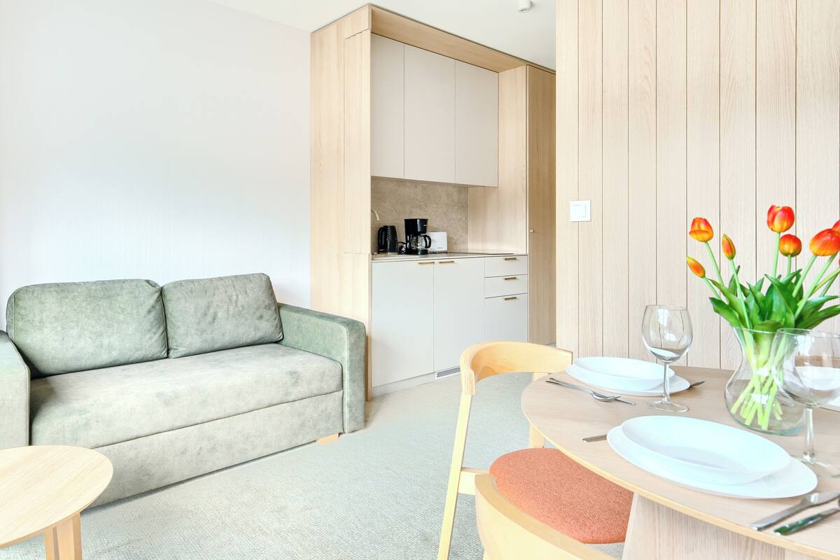 Una habitación de hotel moderna con sofá, una mesa de comedor con sillas, una zona de cocina y un florero con tulipanes.