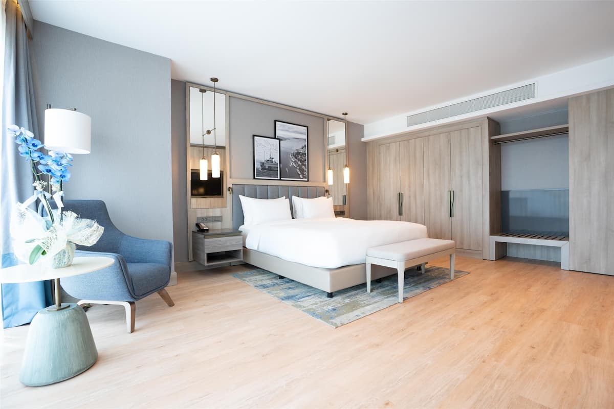 Ein modernes Hotelzimmer mit einem großen Bett, einer Sitzecke mit einem Sessel, einem kleinen Tisch mit einer Vase und einem Kleiderschrank.