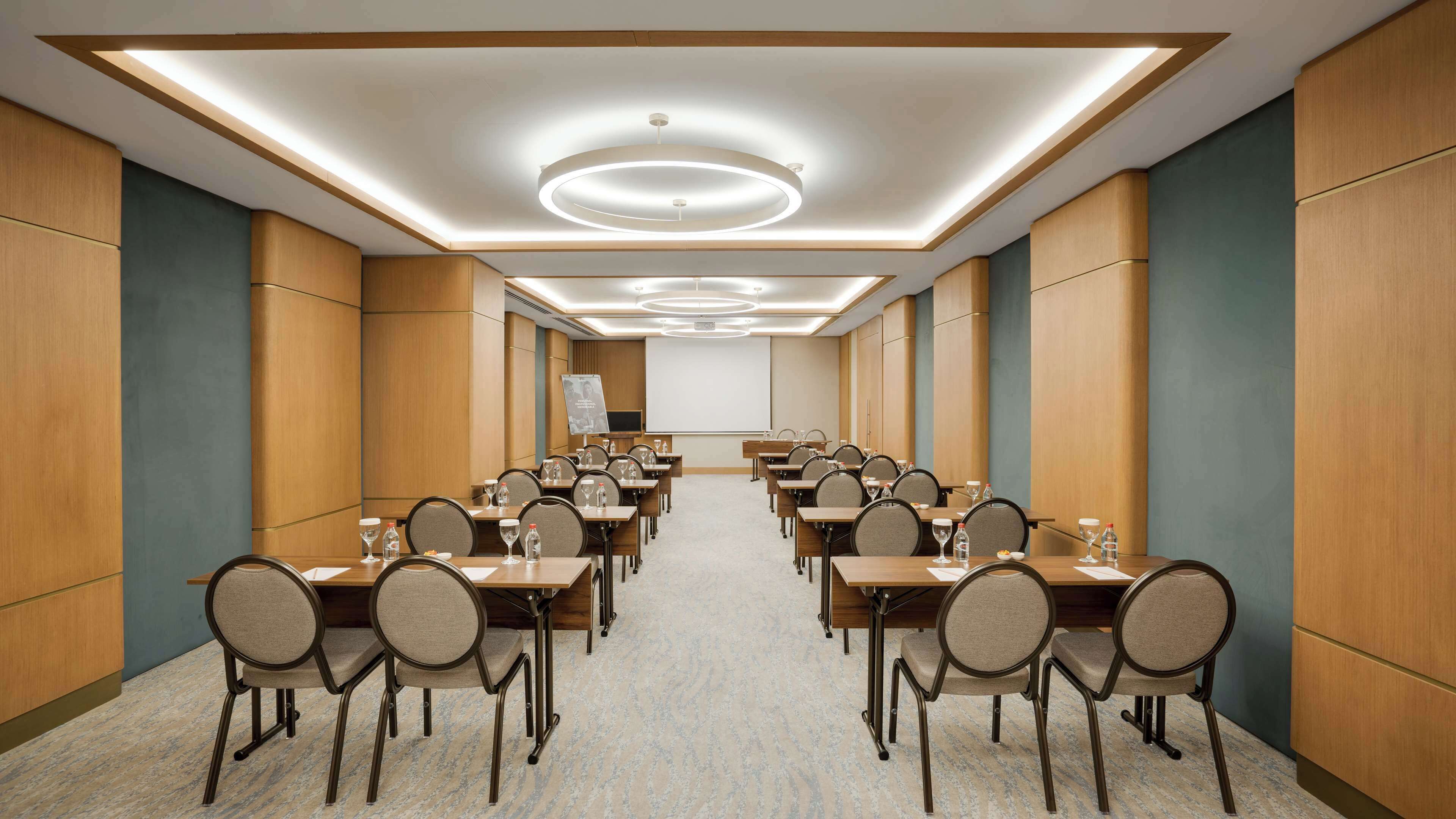 A conference room with long tables, chairs, and a projector screen at the front.
