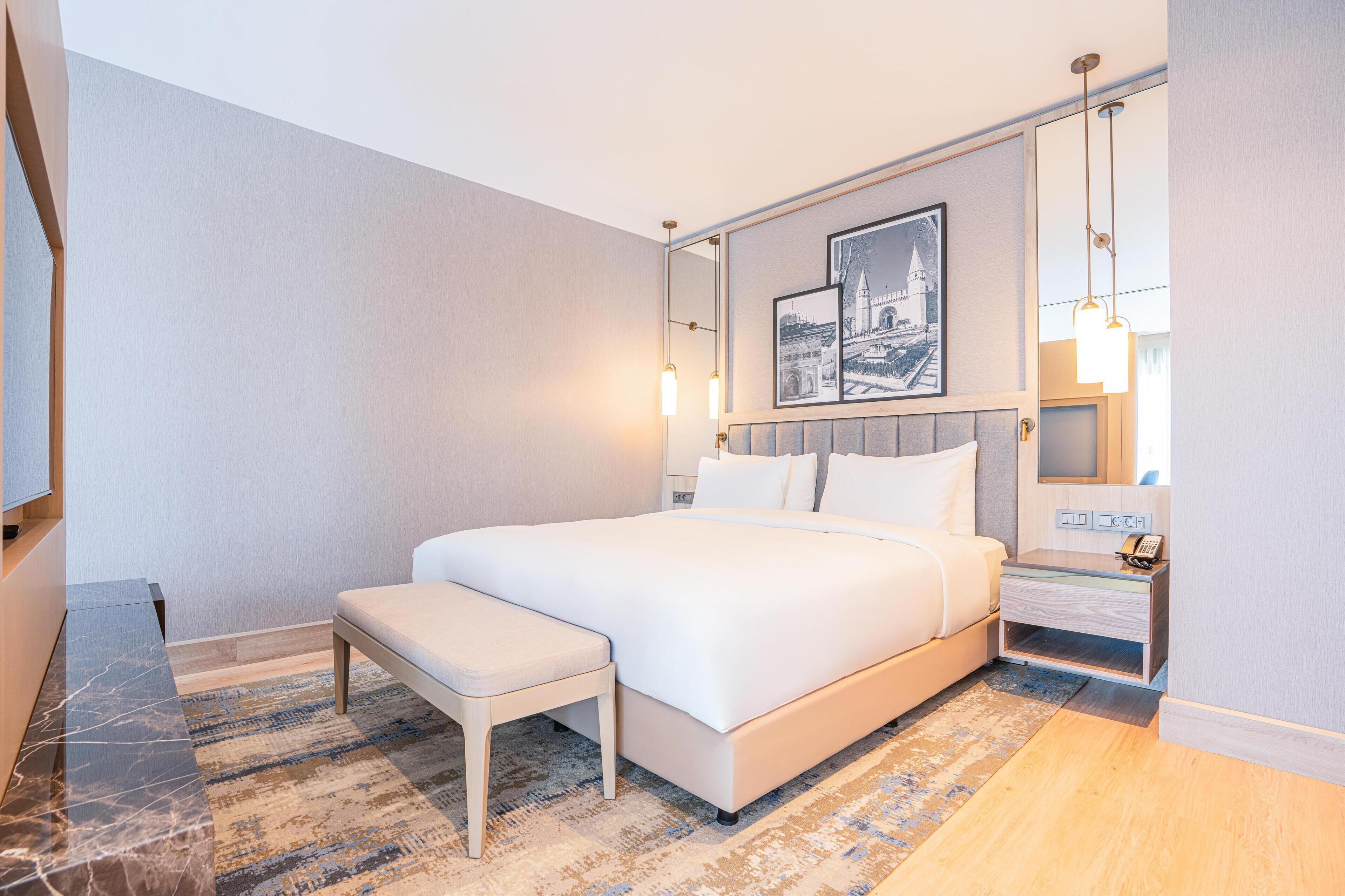 A modern hotel room with a large bed, a bench, a bedside table, and a mirror.