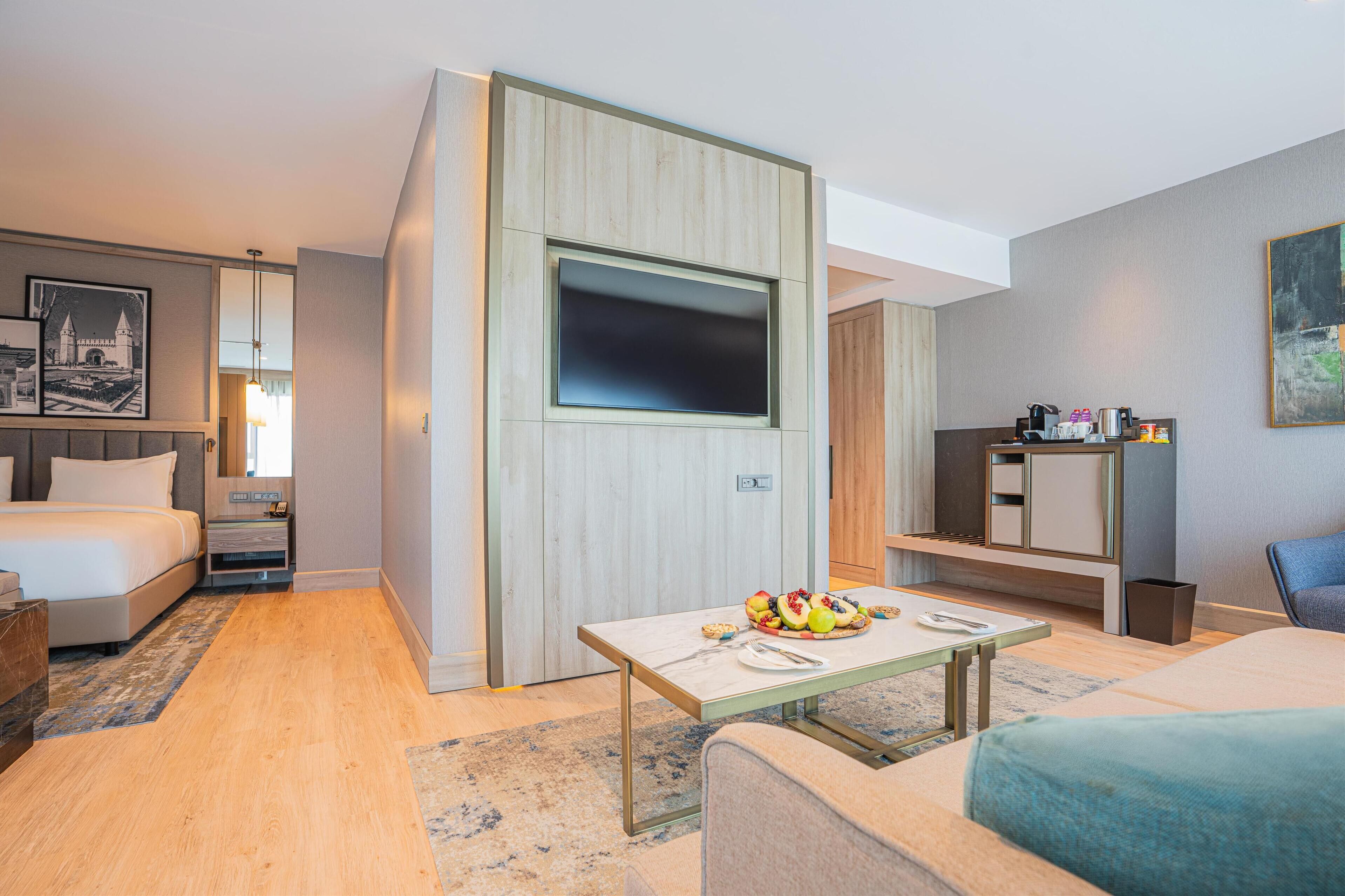A modern hotel room with a bed, a TV mounted on a wooden panel, a small dining table with fruit, a sofa, and a desk with a chair.