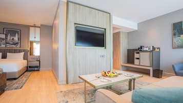 A modern hotel room with a bed, a TV mounted on a wooden panel, a small dining table with fruit, a sofa, and a desk with a chair.