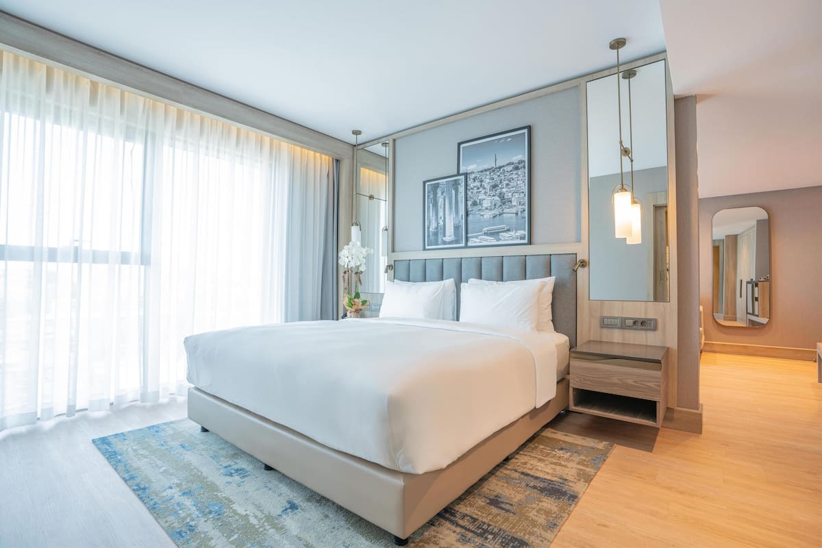 Ein modernes Hotelzimmer mit einem großen Bett, einem Nachttisch, einem Spiegel und Blick auf eine Stadtlandschaft durch das Fenster.